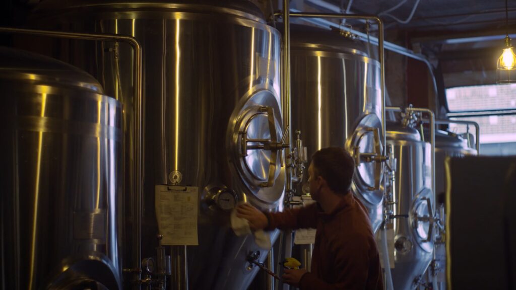 Brewer wipes stainless steel fermentation tanks inside a dimly lit craft brewery production space.
