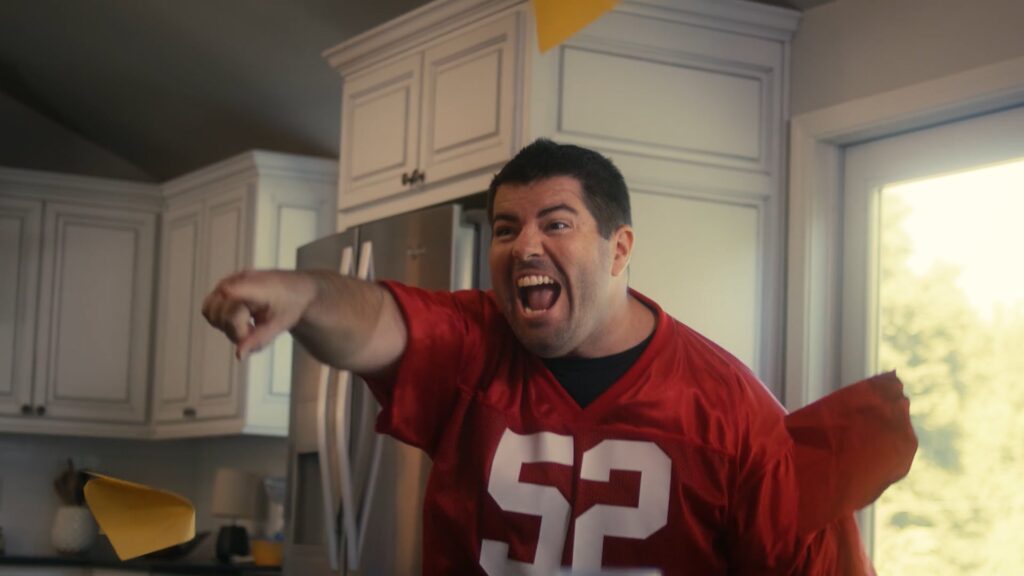 Excited football fan in a red jersey points and cheers in a kitchen during a game day celebration.