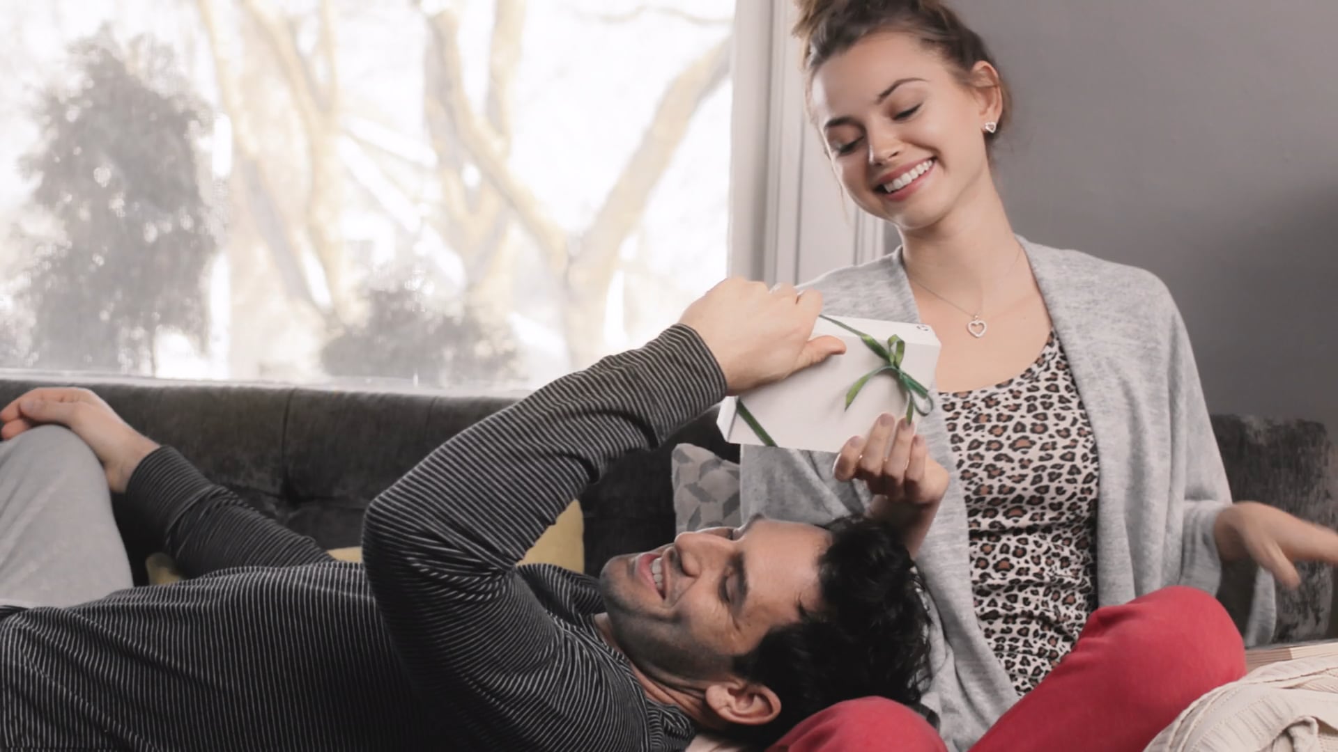 Smiling couple on a couch share a wrapped gift in a cozy living room at home.