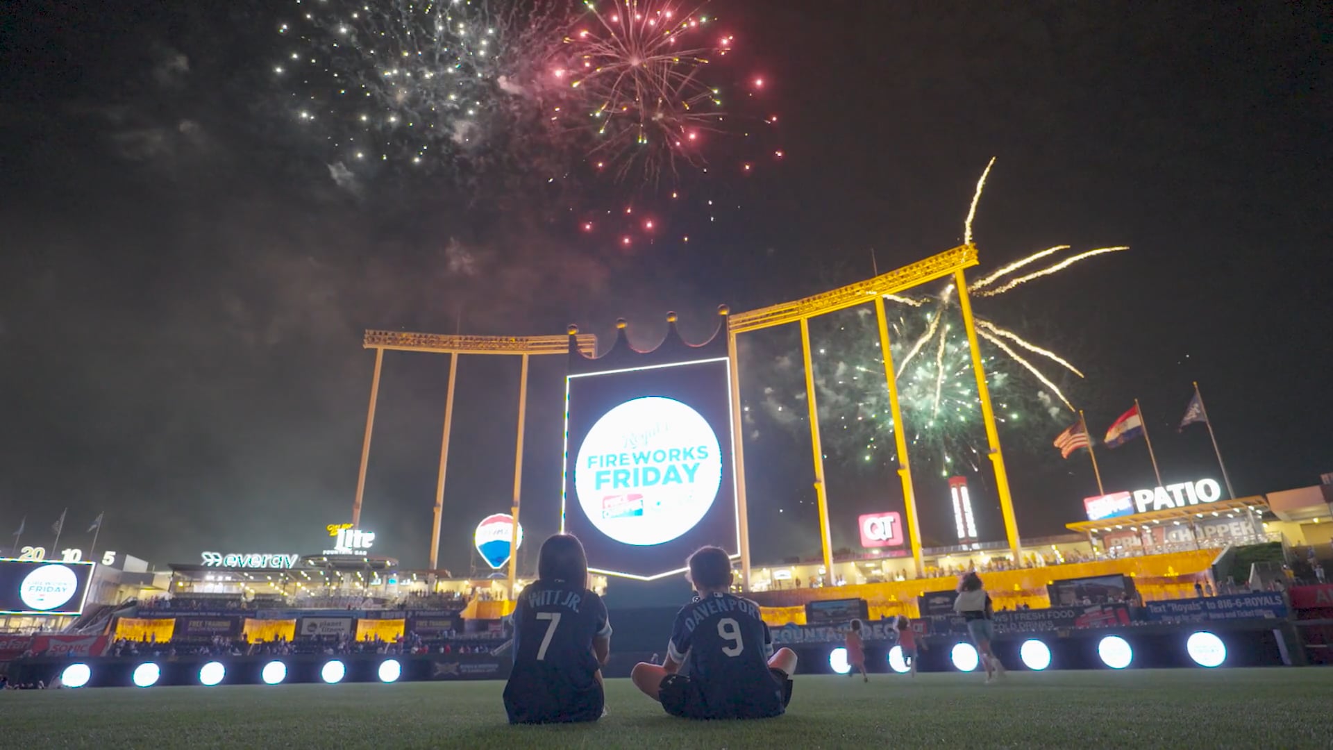 Two fans sit on the field watching fireworks light up a baseball stadium during a summer night game.