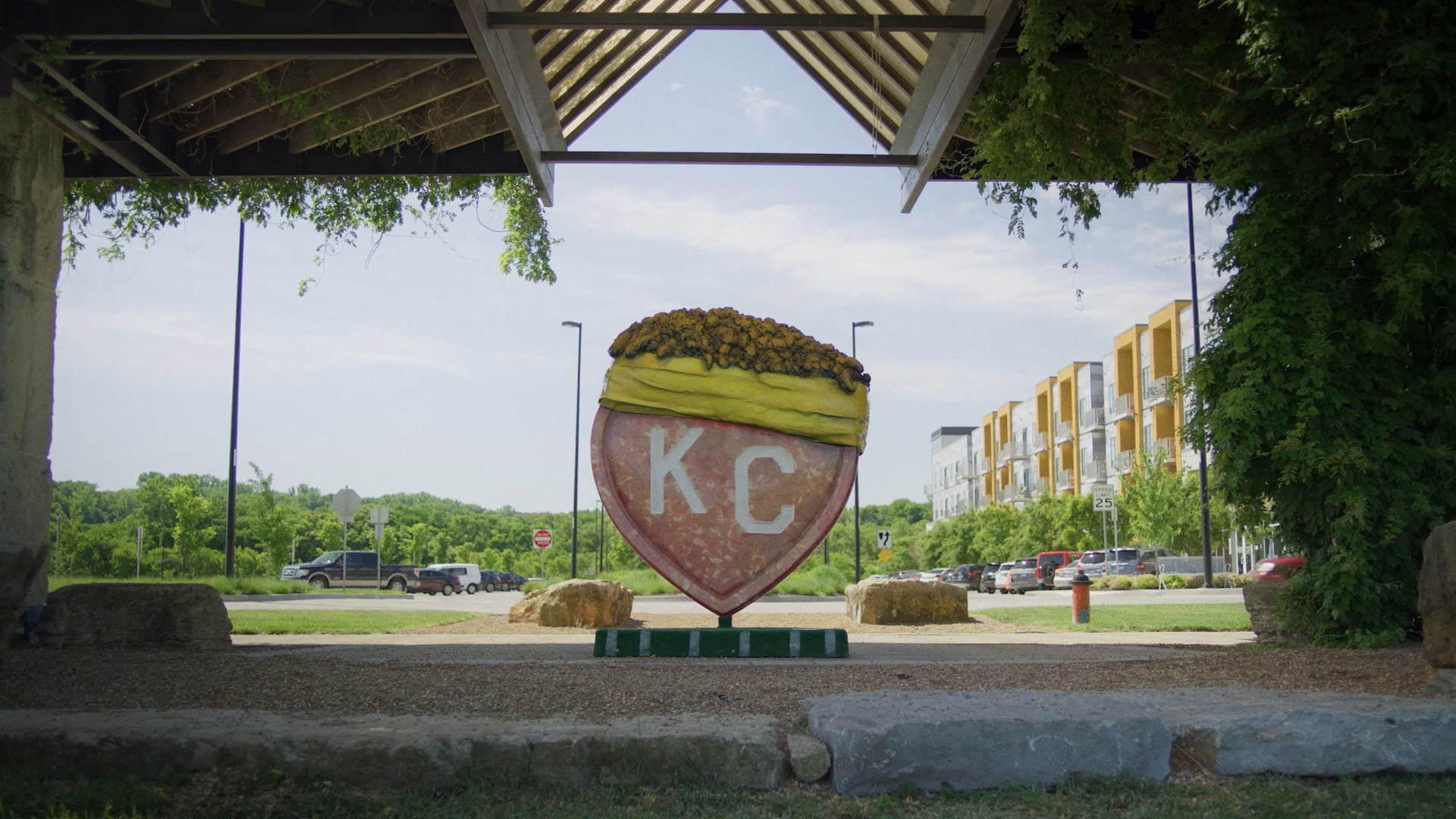 KC heart sculpture under a vine-covered pavilion near apartments and greenery in an outdoor city space.
