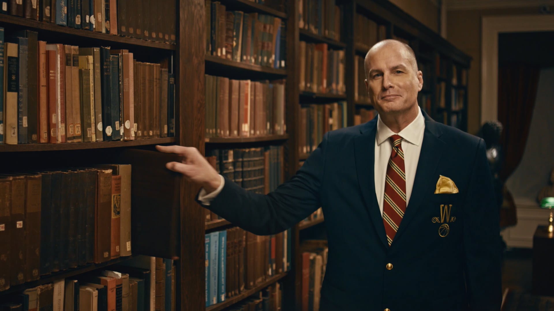 Man in a suit standing by library bookshelves in a formal study, pointing to a book in a classic reading room.