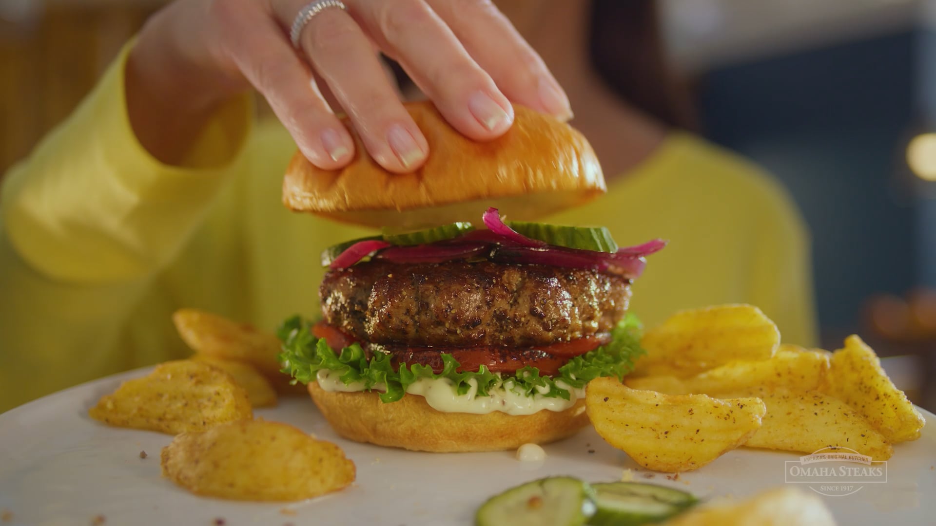 Omaha Steaks burger topped with pickles and red onions on a brioche bun, served with potato chips as a hand places the top bun on the grilled patty.