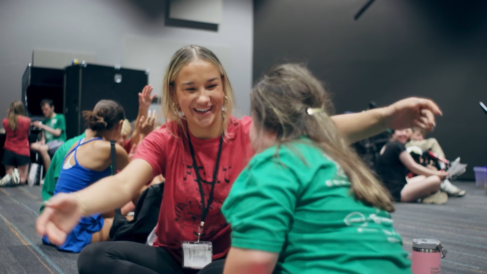 Smiling counselor opens arms to greet a child in a green shirt during an inclusive indoor group activity.