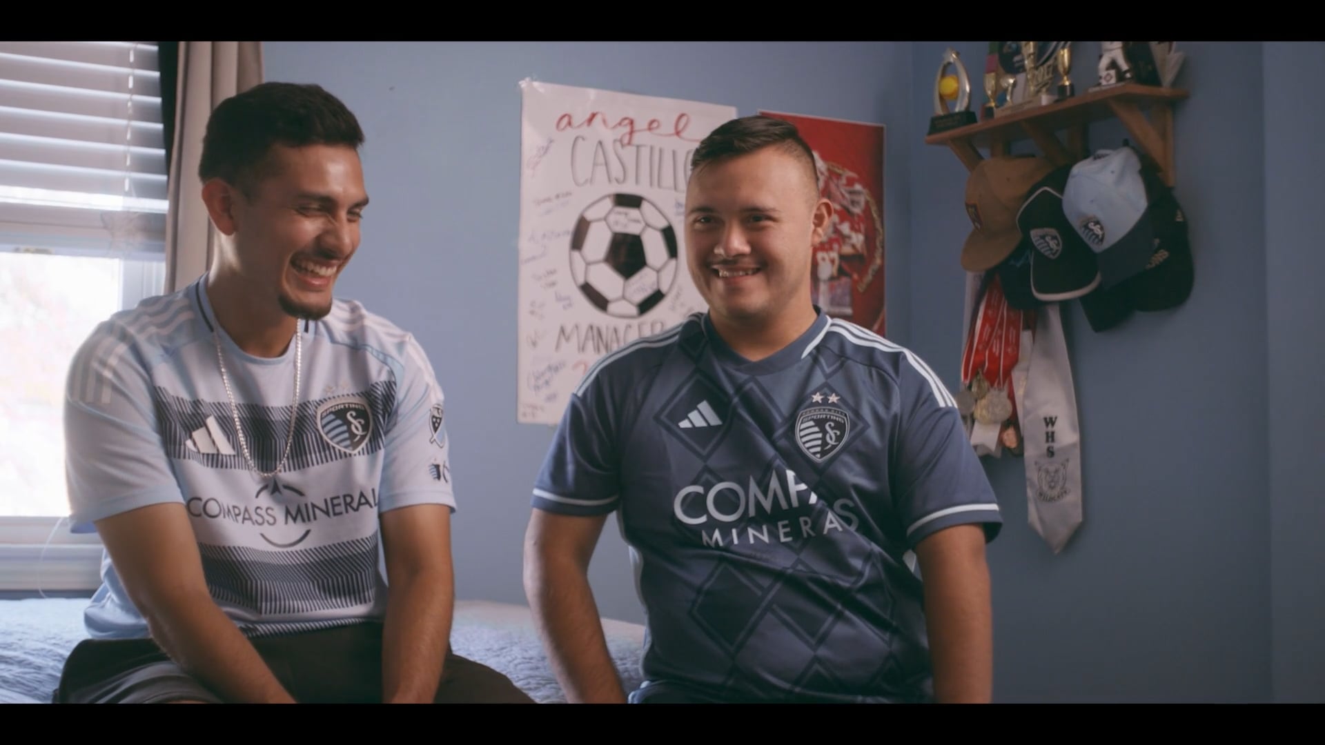 Two smiling soccer fans in Sporting KC jerseys sit in a bedroom with sports memorabilia and team posters.