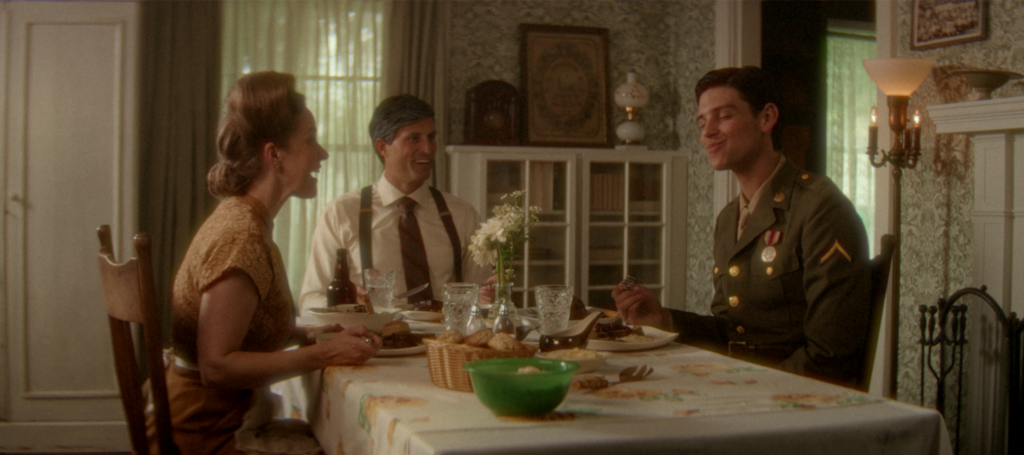 A young soldier in uniform shares a meal and laughs with his parents at their family dining table.