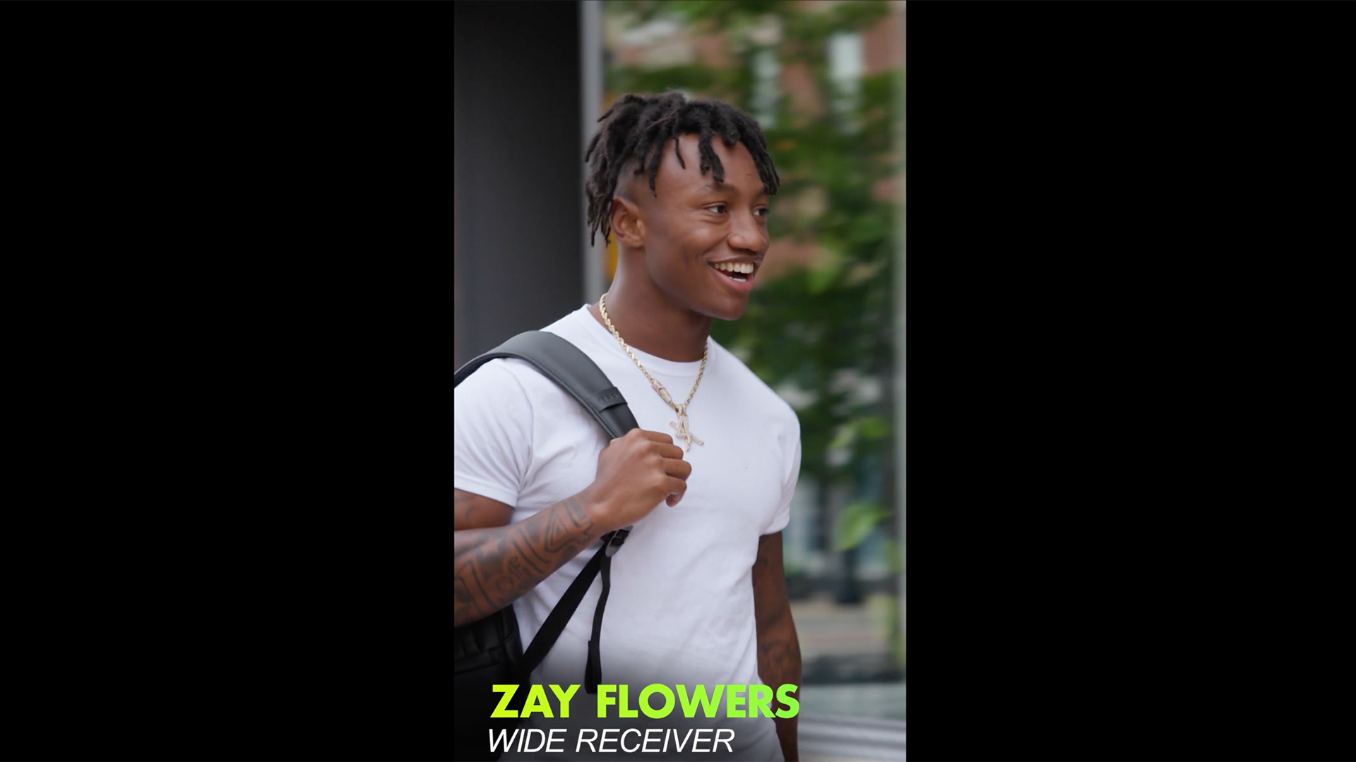 Smiling football wide receiver walking outdoors with a backpack, shown in a vertical player profile graphic.